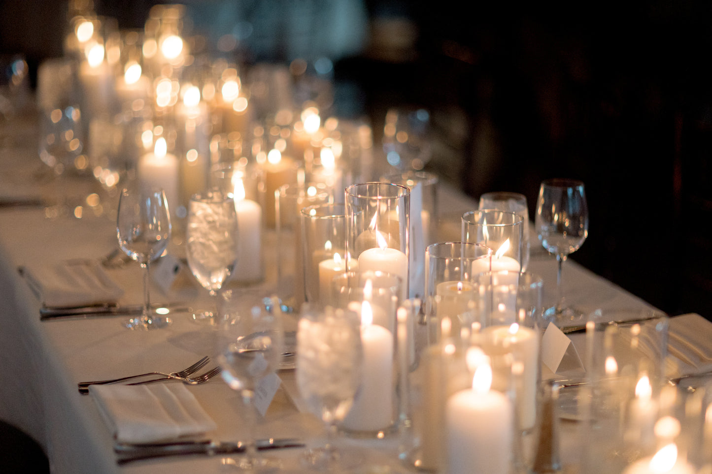 photo of pillar candles in glass hurricanes on a table with white napkins, white tablecloth, and wine glasses