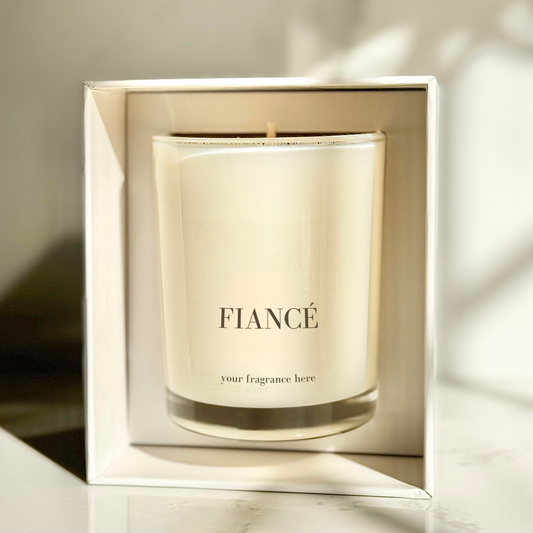 A beige candle with the word 'Fiance' printed on the label, placed on a marble surface with a shadow falling over the background.