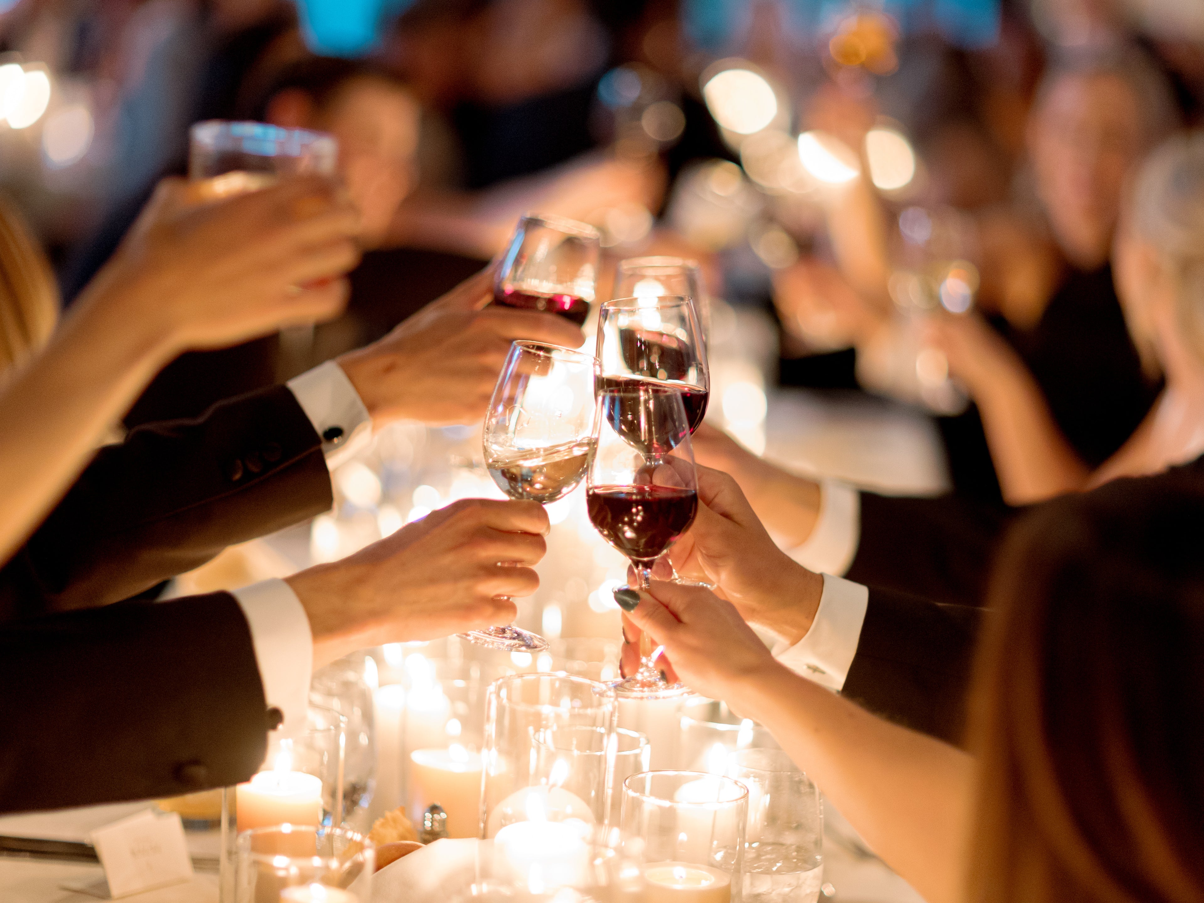 color photo of wine glasses clinking at a wedding with a candlelit table below the glasses
