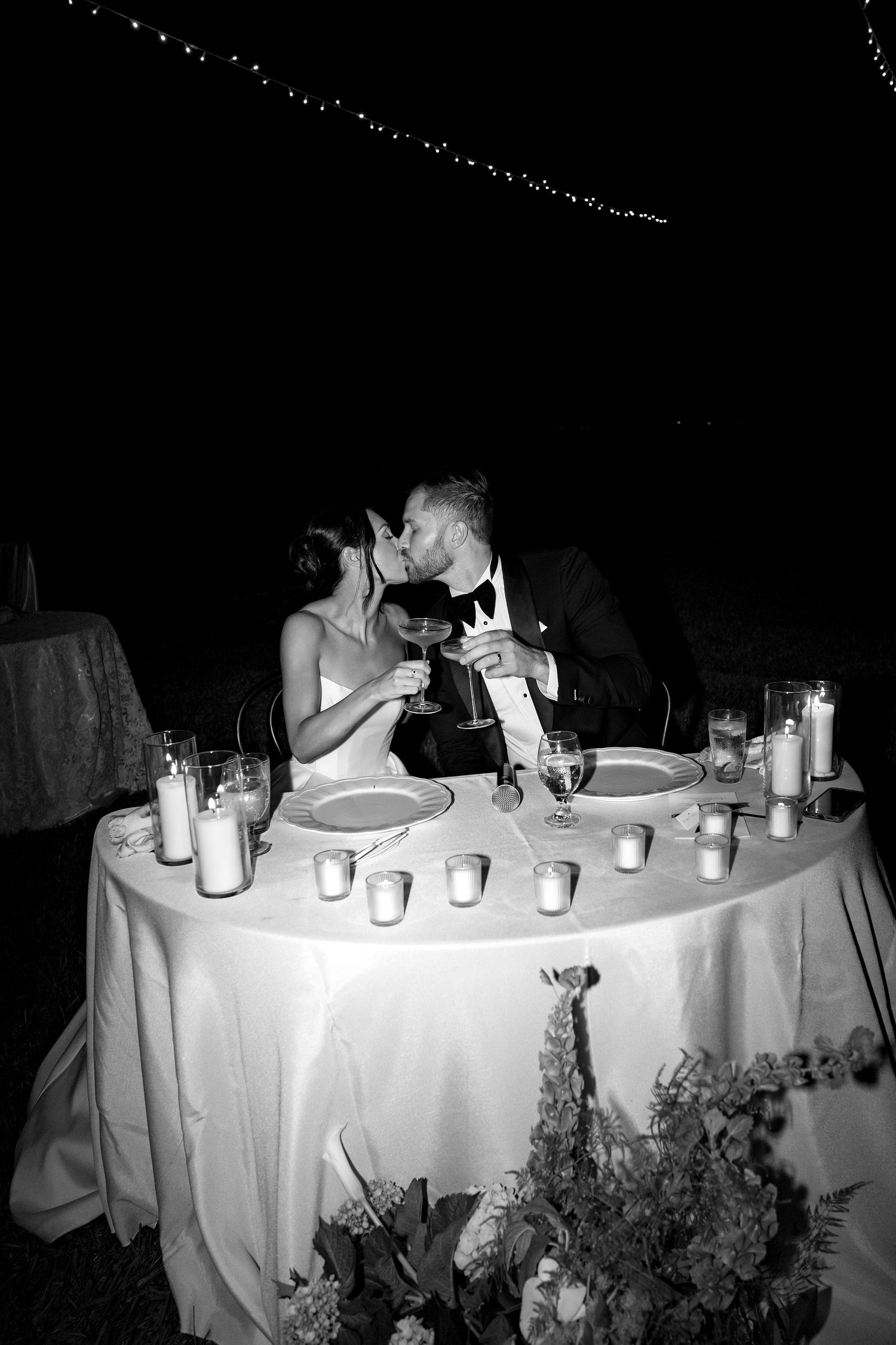 Black and white photo of a couple kissing at a decorated table with candles and glasses.