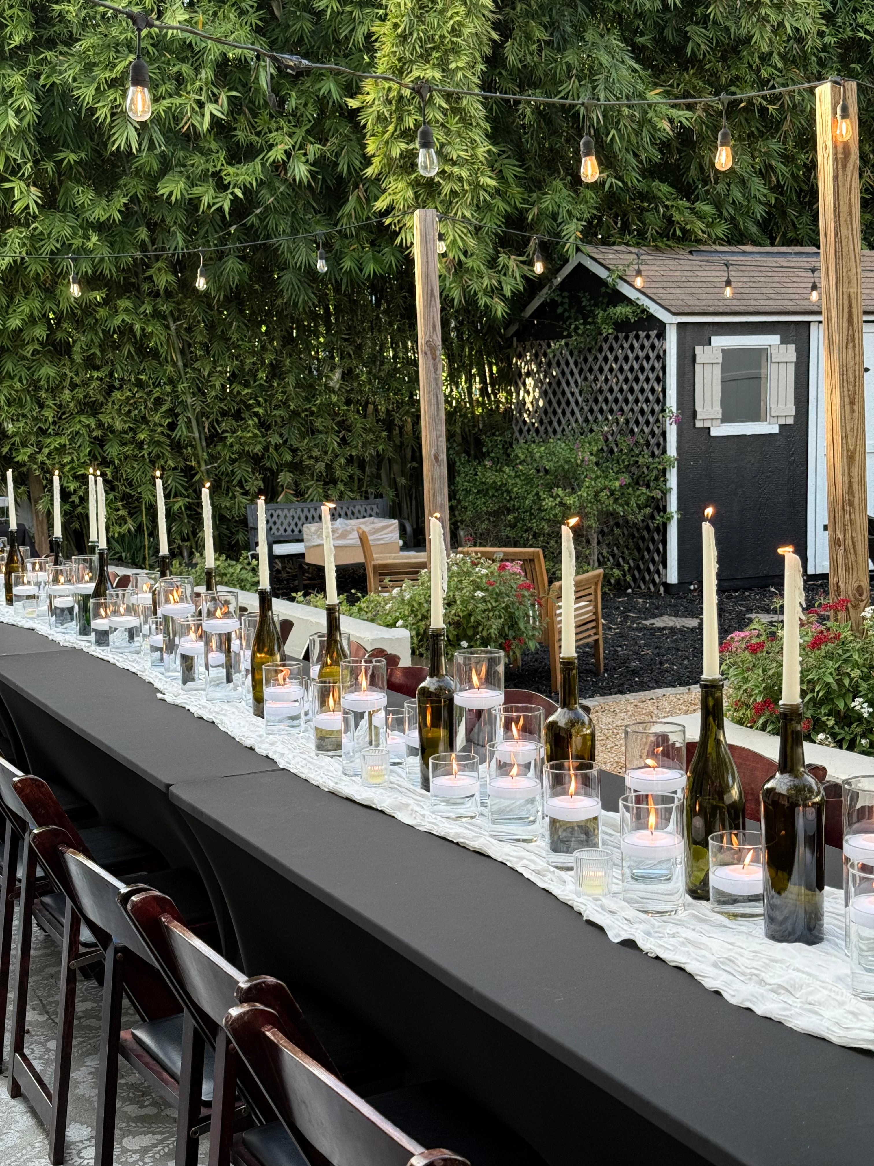 Outdoor table setting with wine bottles, glasses, and candles under string lights.